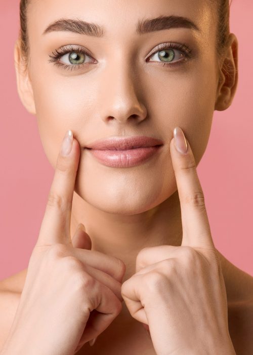 Natural beauty. Young woman applying lip balm, holding fingers near lips, pink background