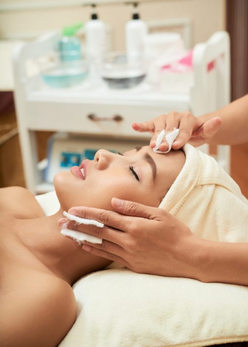 Close-up shot of attractive young woman lying on treatment table with closed eyes and receiving procedure, interior of modern facial room on background